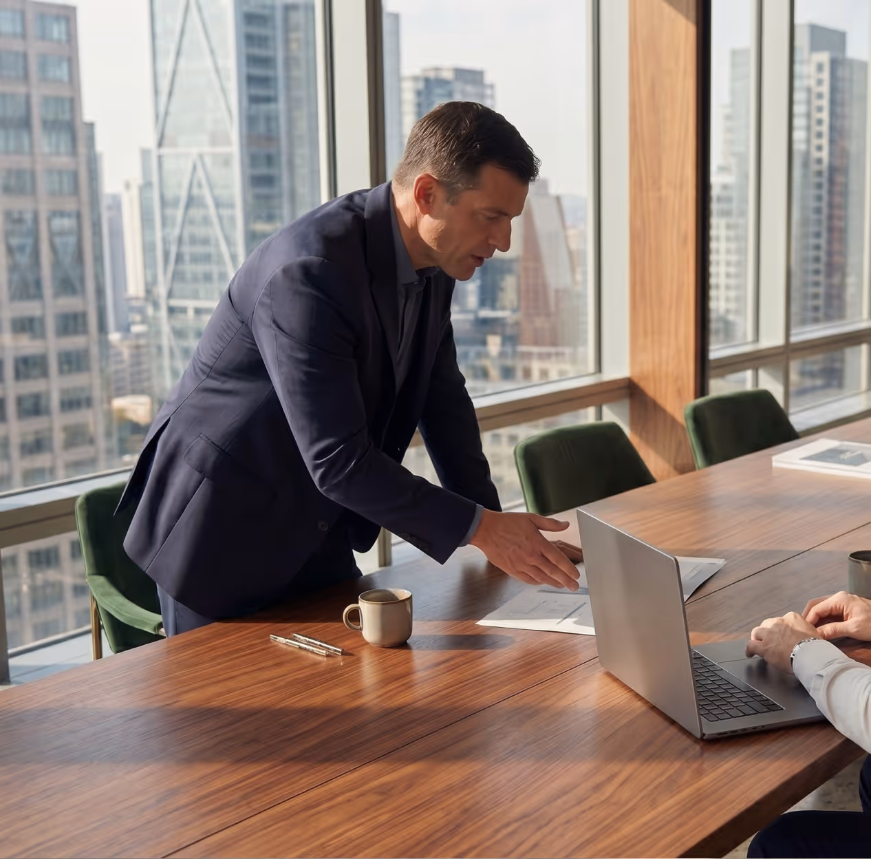 Man in suit leaning over conference table discussing papers with colleague using laptop.