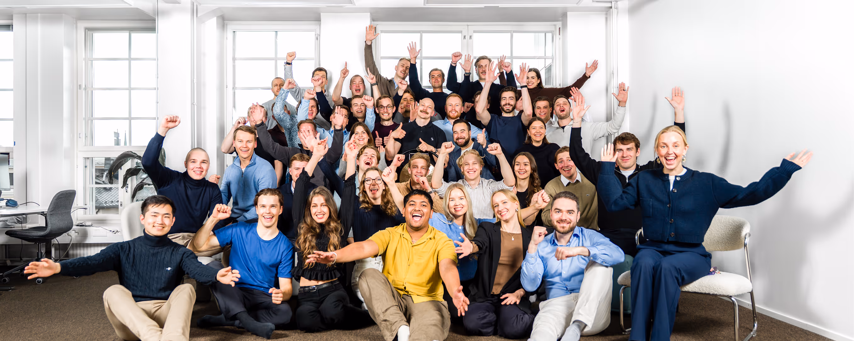 Large group of diverse young adults smiling and raising hands in bright office space.