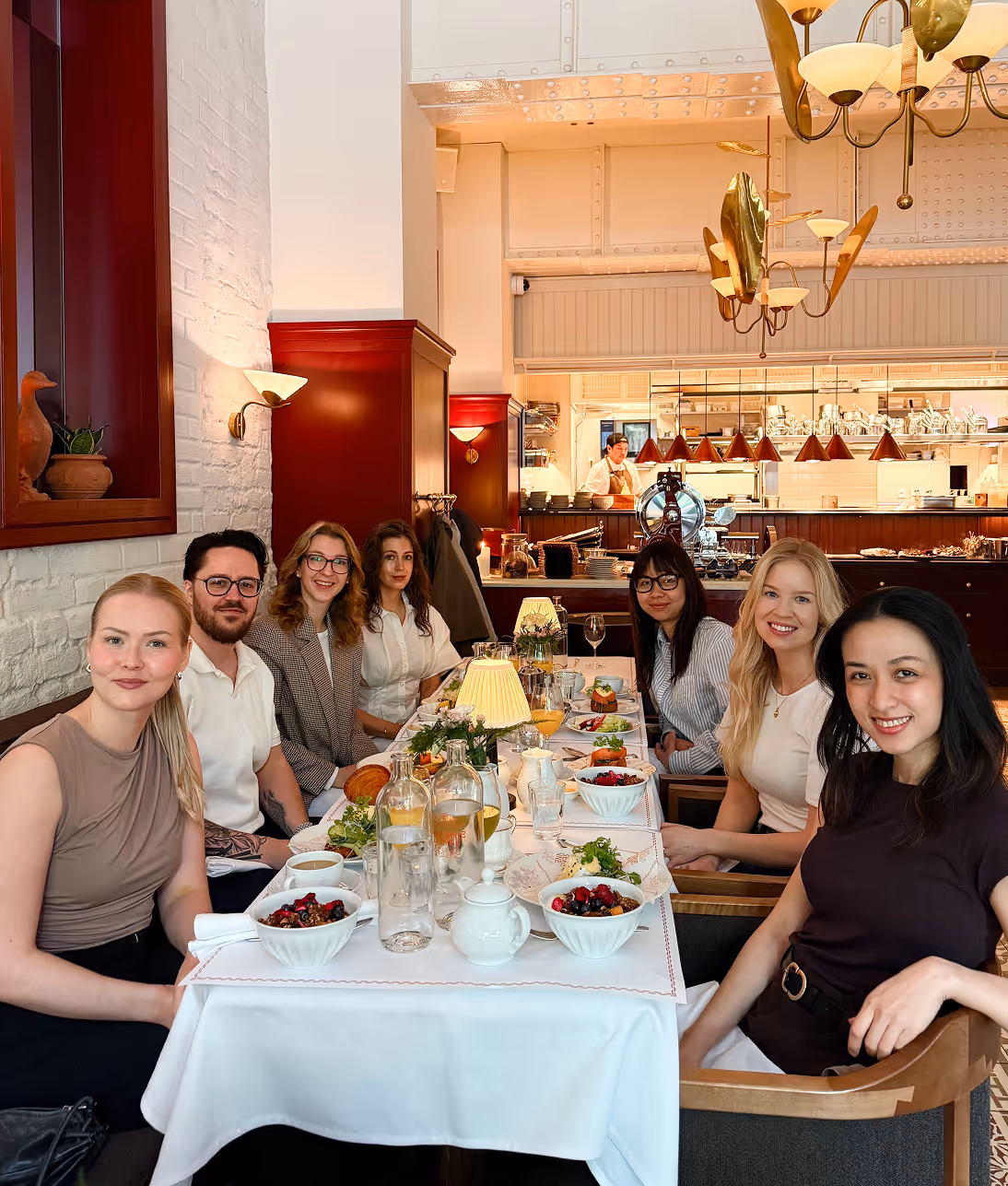 Group of seven adults smiling and seated at a long table with food in a warmly lit restaurant.