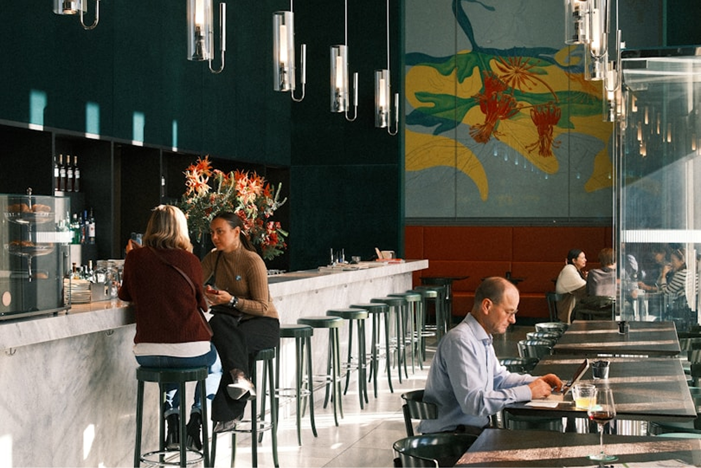 Deux femmes parlent au comptoir tandis qu'un homme travaille sur un ordinateur dans un café moderne.