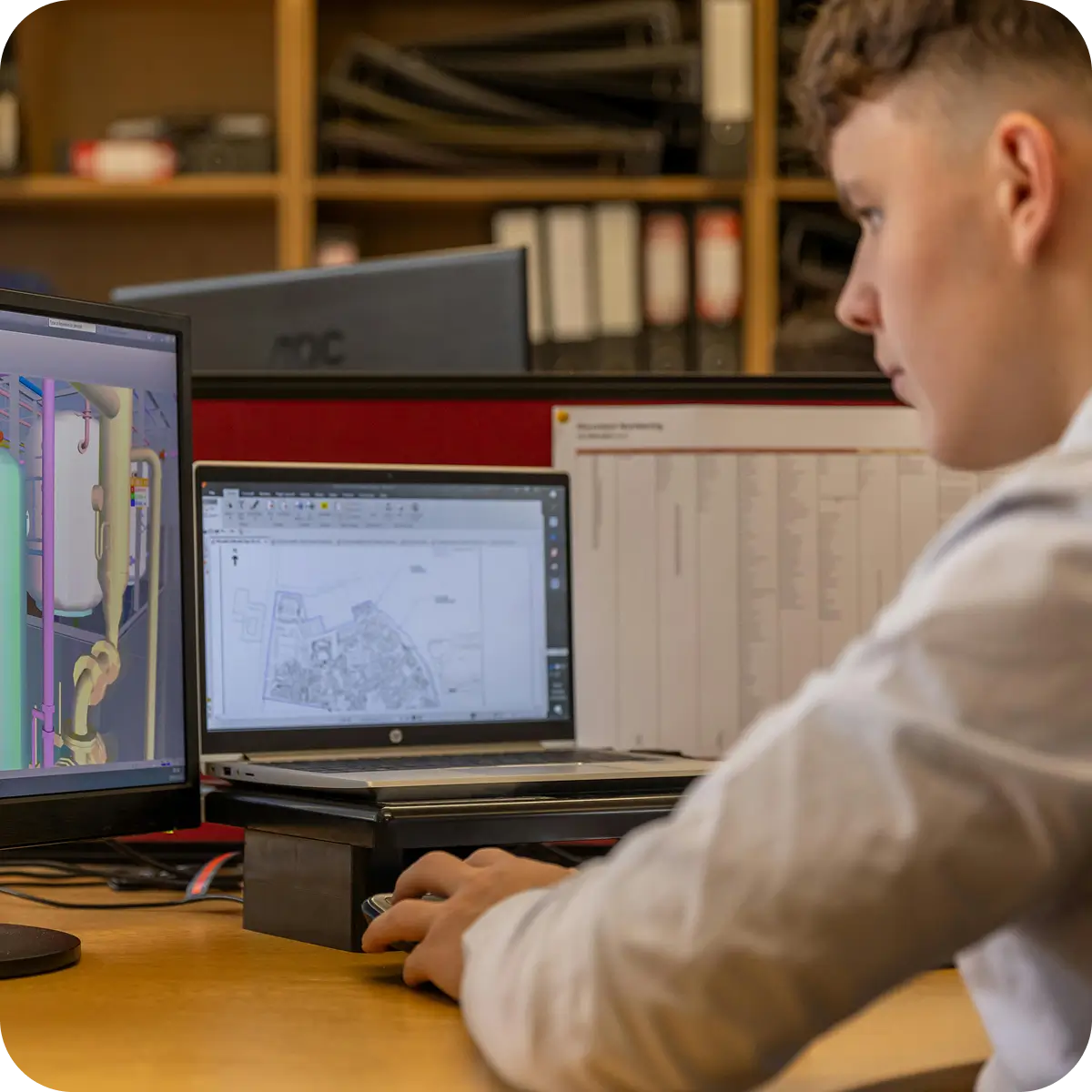 Jeune homme concentré travaillant sur ordinateur avec un logiciel de modélisation 3D et un plan technique à l'écran.