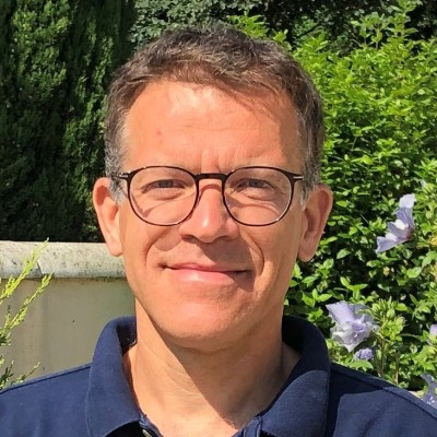Homme portant des lunettes souriant devant des arbustes et des fleurs violettes au soleil.