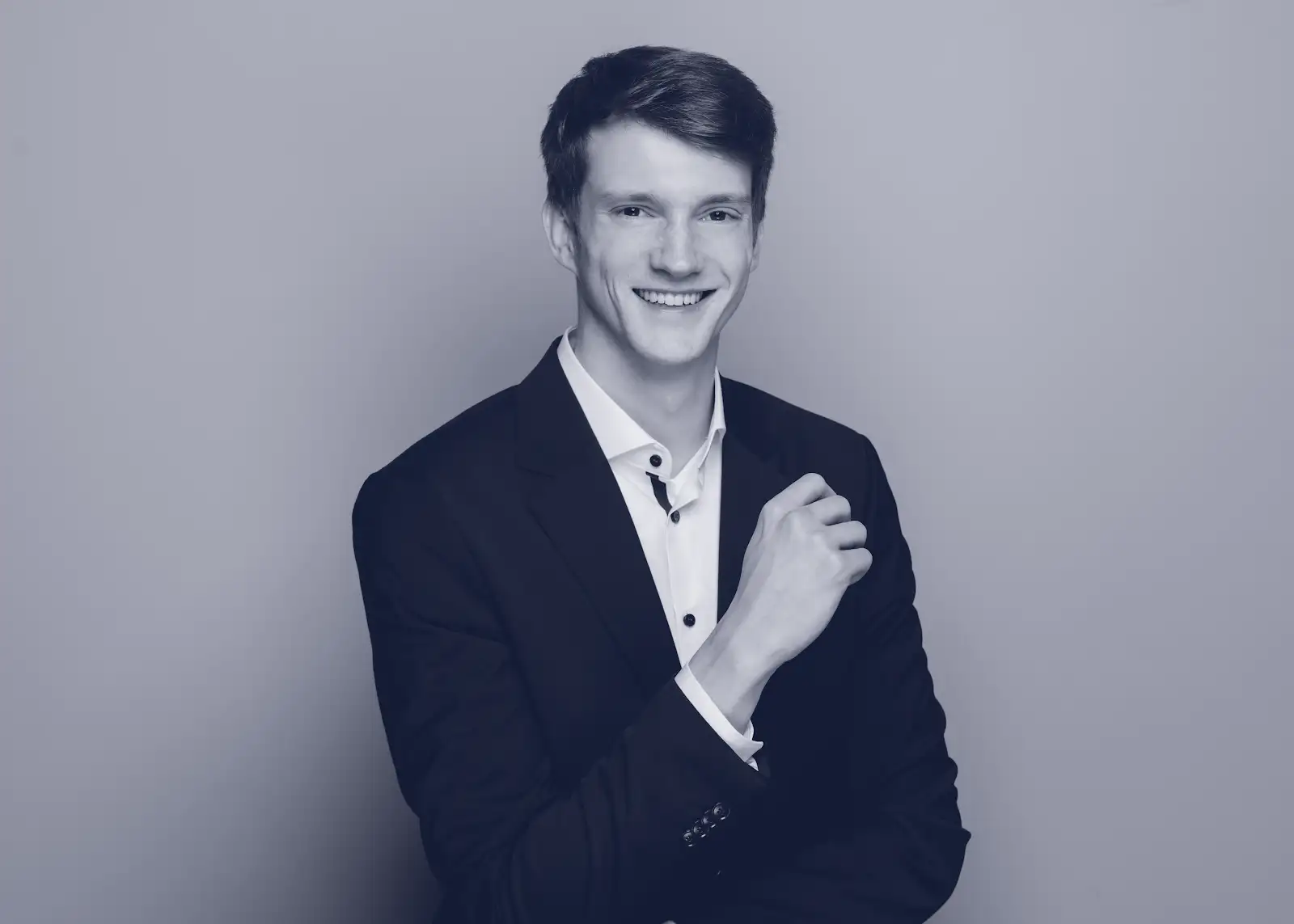 Young man in a dark suit smiling and adjusting his cufflink against a plain background.
