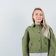 Smiling woman with long blonde hair wearing a green jacket over a gray top, standing against a plain light gray background.