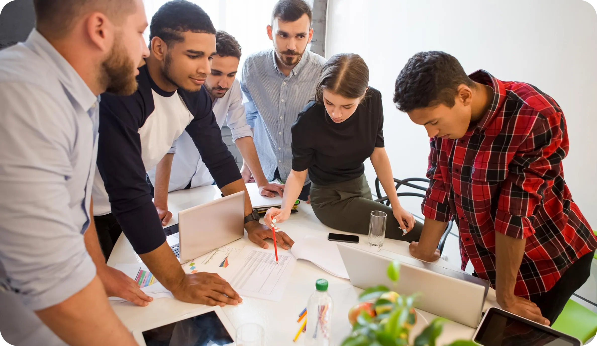 Group of six young professionals gathered around a table discussing documents and laptops in an office.