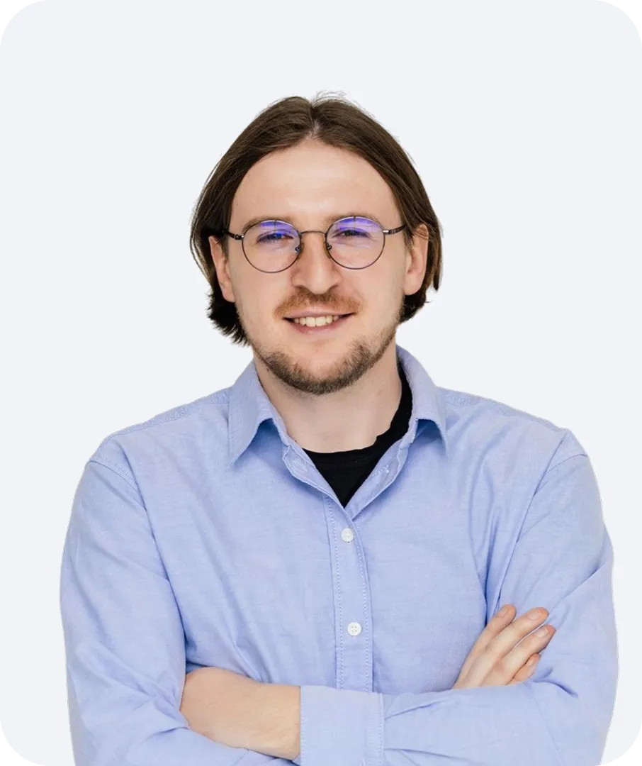 Smiling man with medium-length brown hair and glasses, wearing a light blue button-up shirt and black undershirt, arms crossed.