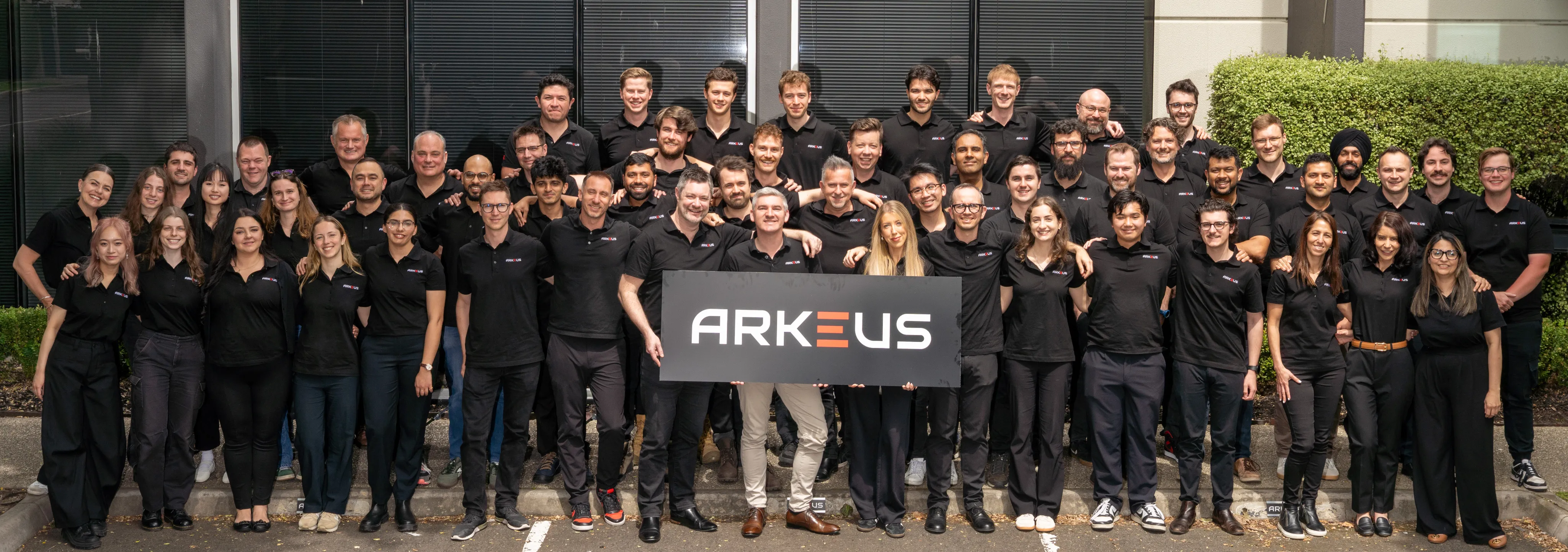 Large group of smiling people wearing black ARKEUS polo shirts posing outside a building, holding a sign with the ARKEUS logo.