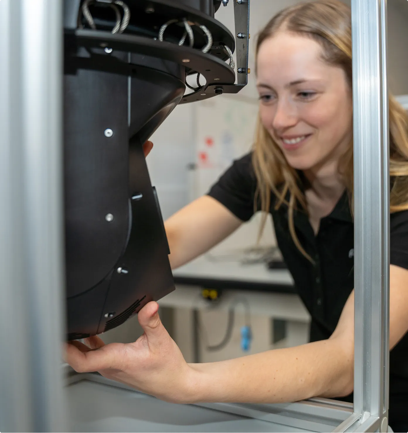 Woman adjusting a black mechanical or electronic device inside a metal-framed enclosure.