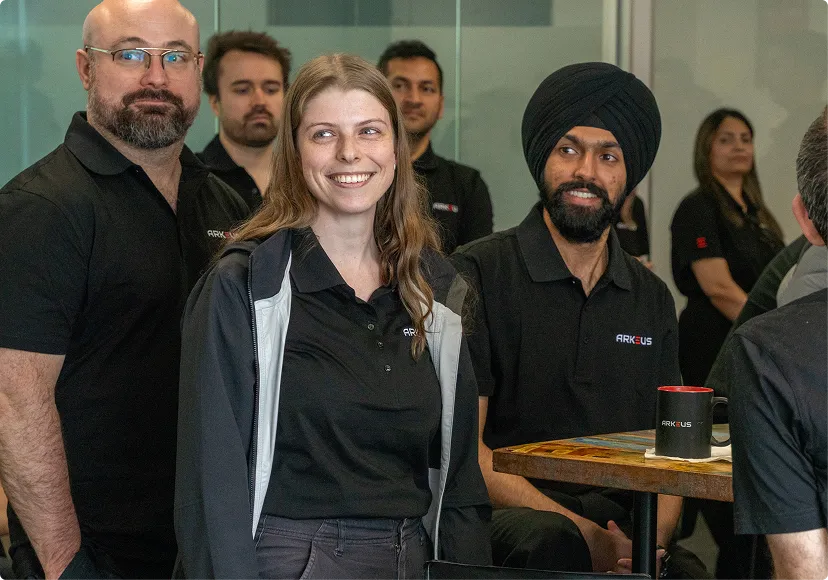 Group of diverse coworkers wearing black ARKEUS shirts smiling and sitting around a table with an ARKEUS coffee mug.