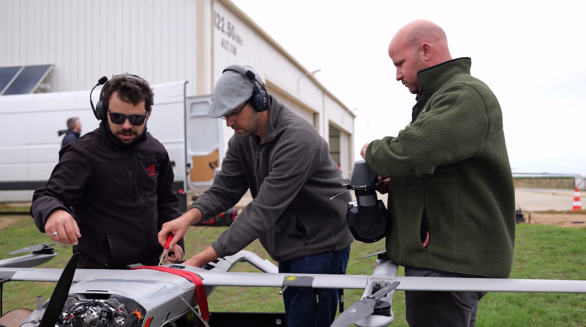 Three men wearing headsets working on a large drone outdoors near a hangar.