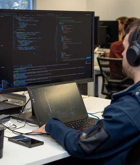 Person wearing headphones working on code displayed on a large monitor and laptop in an office setting.