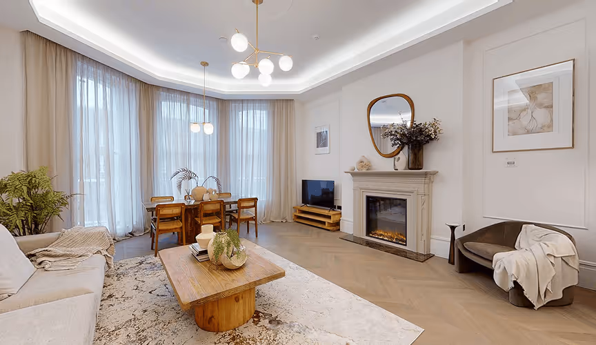 Bright living room with large windows, a dining table, fireplace, sofa, armchair, and a wooden coffee table on a rug.