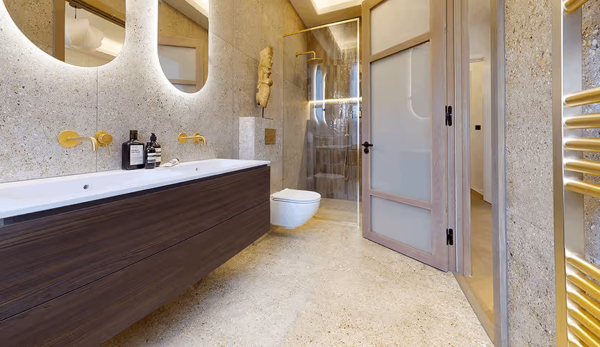 Modern bathroom with double sinks, gold fixtures, a wall-mounted toilet, and a glass shower enclosure.