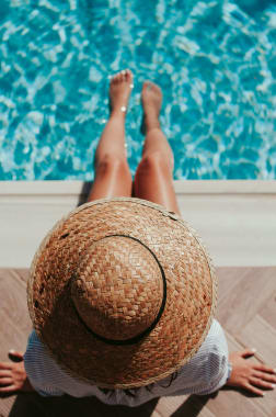 Person wearing a large straw hat sitting by a blue swimming pool with legs extended toward the water.