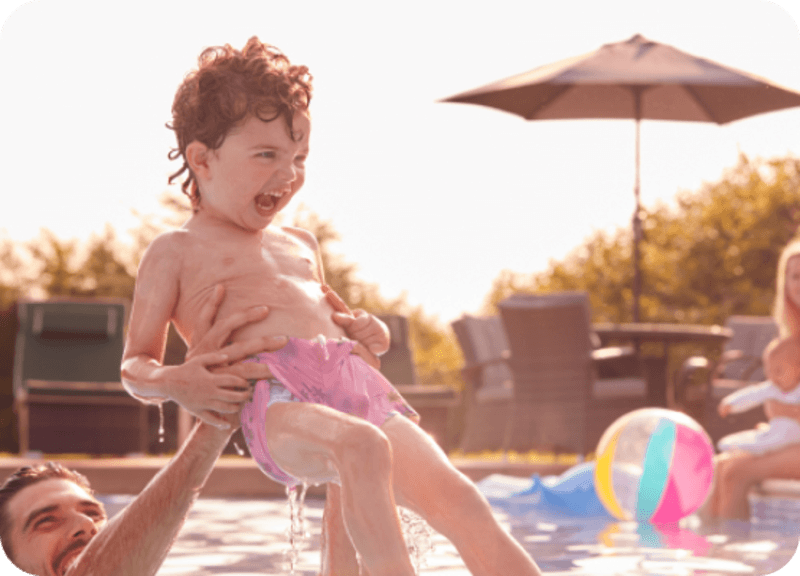 Man lifting joyful wet toddler wearing pink swim shorts above a pool on a sunny day.