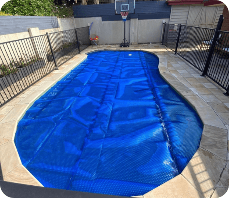 Outdoor pool covered with a blue safety cover surrounded by beige stone tiles and black metal fencing.