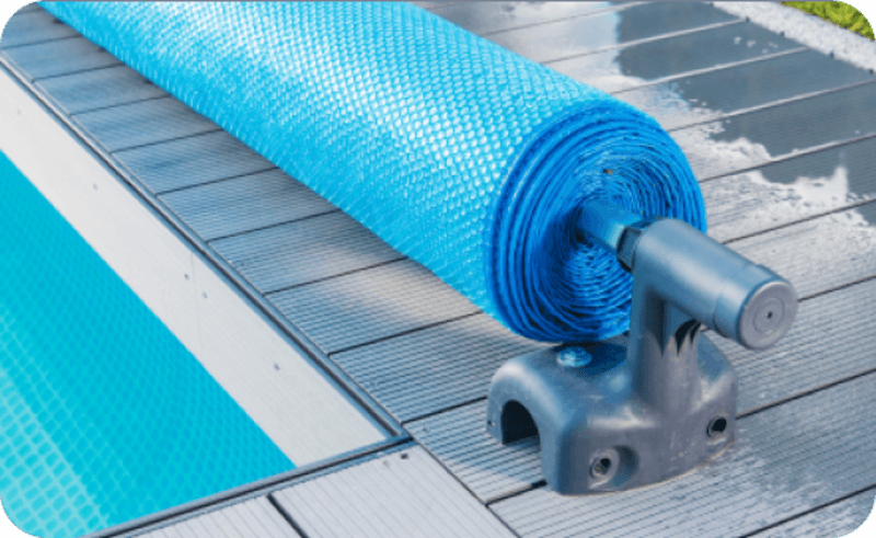 Blue pool cover rolled on a white plastic reel next to a pool on a wooden deck.