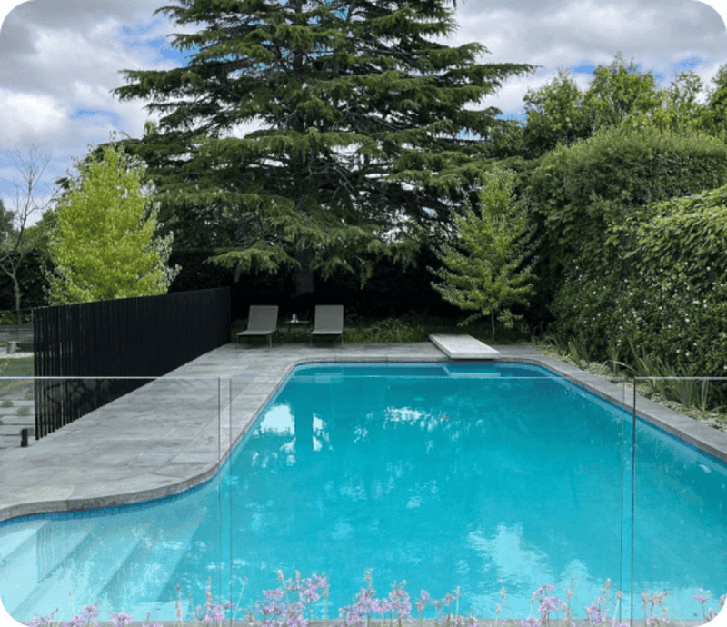 Clear blue rectangular pool with a diving board and two lounge chairs surrounded by trees and shrubs.