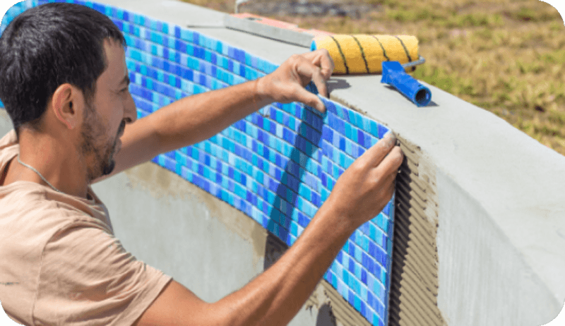 Man installing blue mosaic tiles on the curved edge of an outdoor pool under construction.