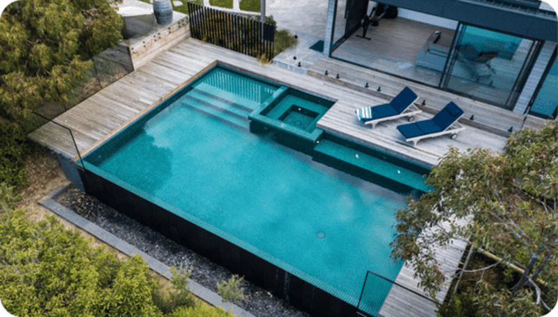 Modern rectangular swimming pool with attached spa and two blue lounge chairs on wooden deck.