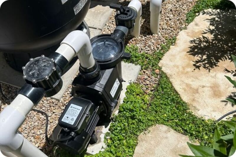 Outdoor pool pump system with pipes and valves surrounded by small stones and stepping stones amid green ground cover.