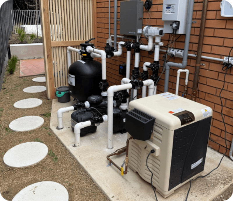 Outdoor pool pump system with black filter, white PVC pipes, and beige heater unit near brick wall.