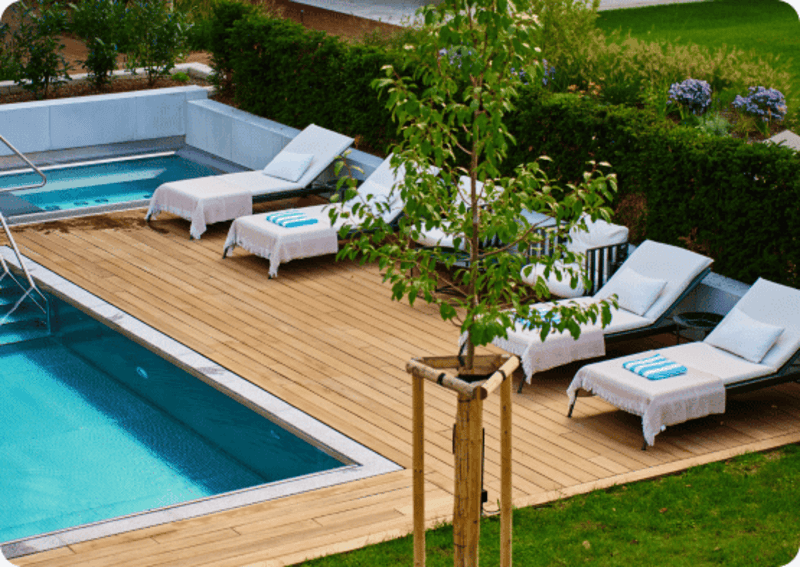 Wooden deck with white lounge chairs around a swimming pool and small tree in foreground.