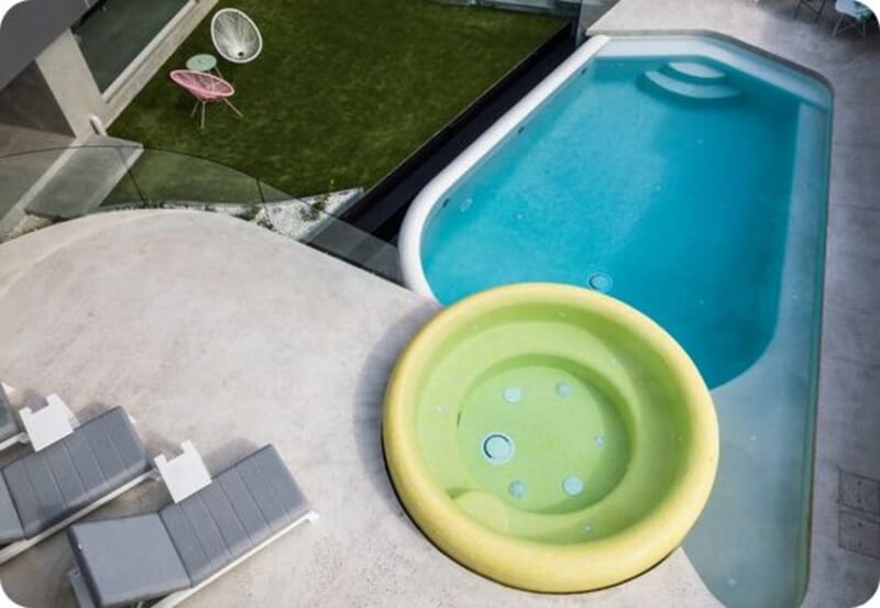 Modern outdoor area with an oval swimming pool, round hot tub, lounge chairs, and green lawn with chairs.