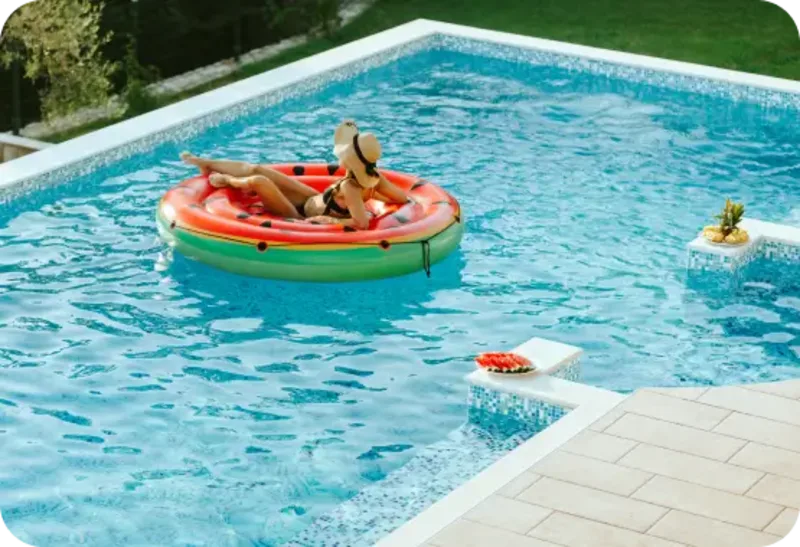 Woman lounging on a watermelon-shaped float in a clear blue pool with fruit on the edge.