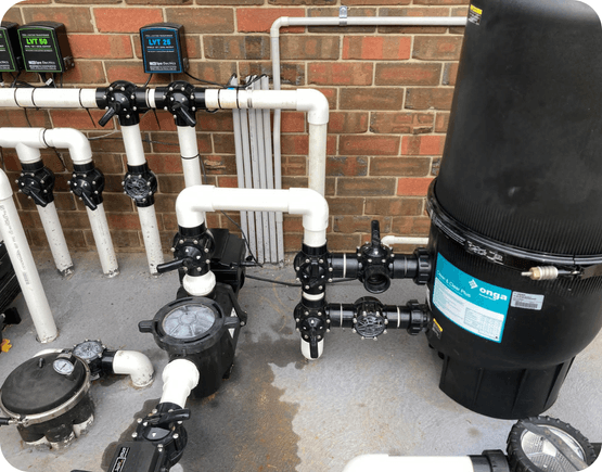 Swimming pool filter system with black and white pipes, gauges, valves, and a large Onga filter against a brick wall.