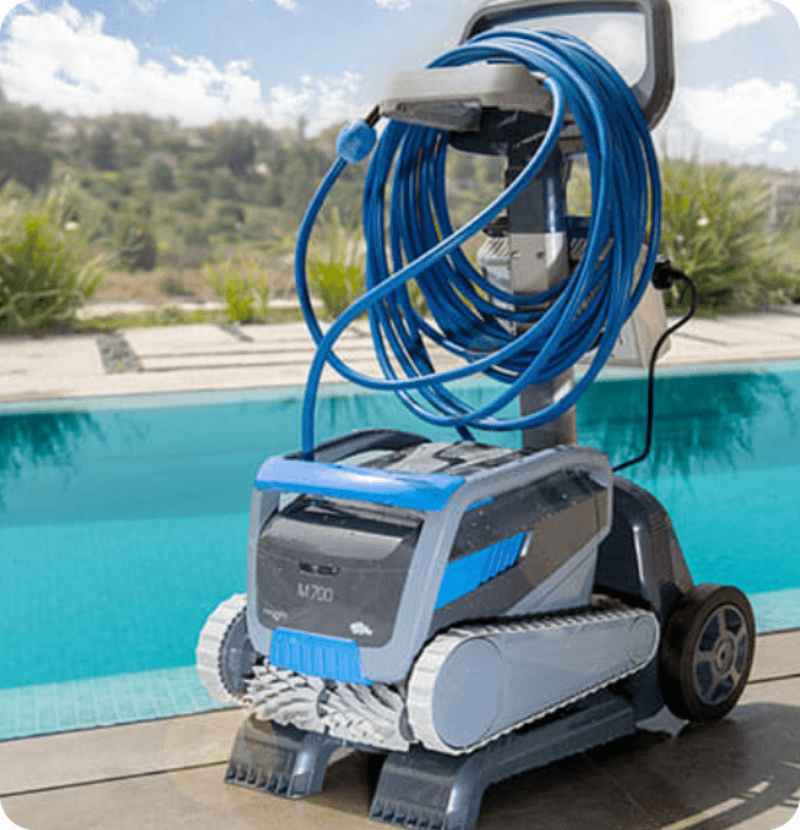 Blue and gray pool cleaning robot with coiled hose beside a backyard swimming pool.