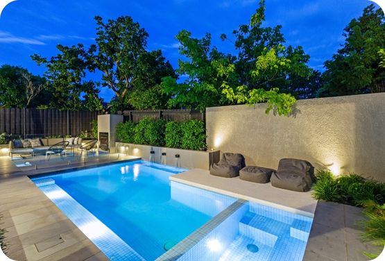 Lit outdoor swimming pool and hot tub with seating area at dusk surrounded by trees and plants.