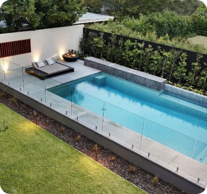 Modern backyard with rectangular pool, glass fence, lounge chairs, and green lawn.