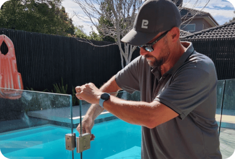 Man in cap and sunglasses adjusting a glass pool fence near a swimming pool outdoors.