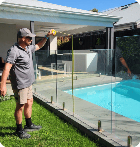 Man measuring height of glass pool fence beside backyard swimming pool on sunny day.
