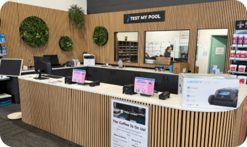 Store counter with wood paneling, computer screens, pool testing sign, and a boxed robotic pool cleaner on display.
