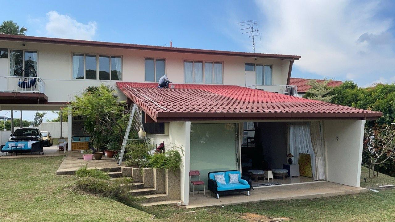 Person painting red roof tiles of a white two-story house with outdoor patio and lawn.