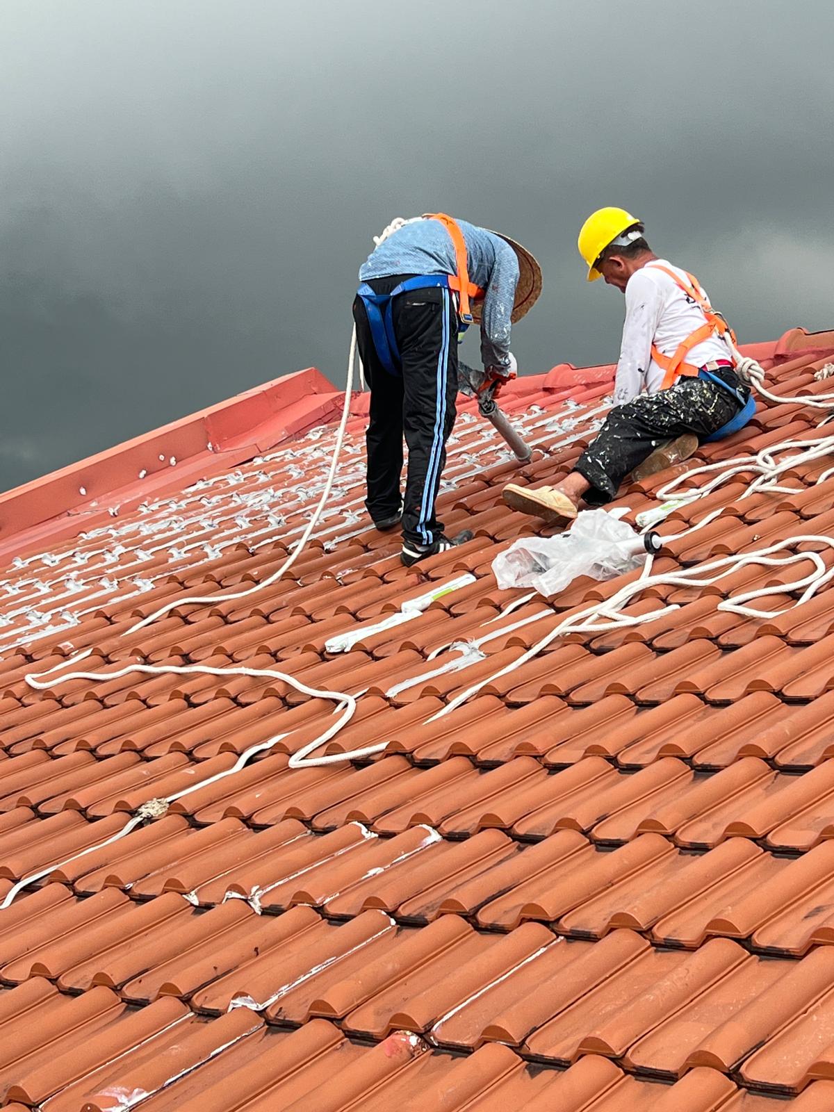Two workers in safety harnesses applying sealant on a tiled roof beneath a cloudy sky.