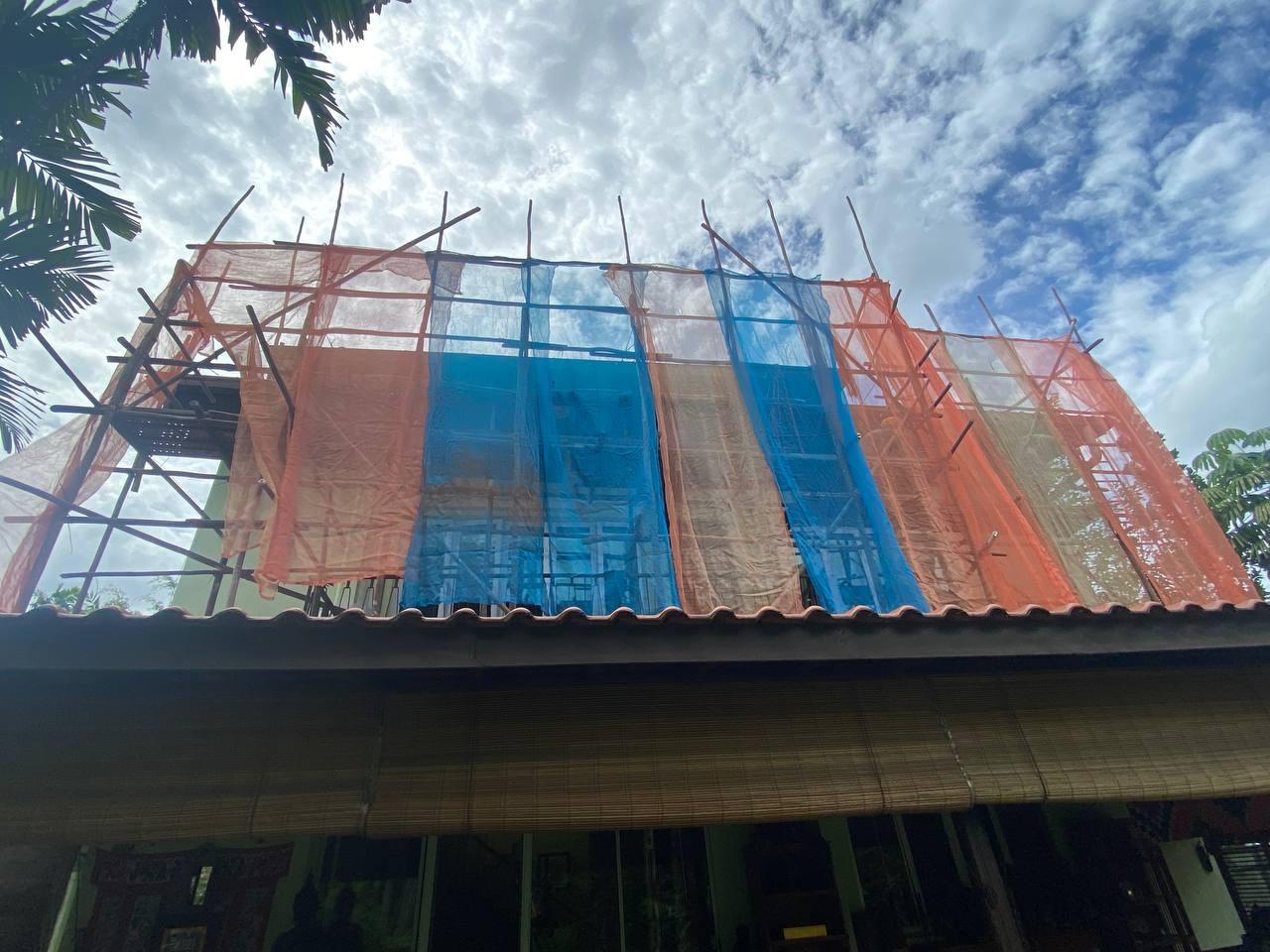 Building under construction with scaffolding covered in orange, blue, and beige safety nets against a partly cloudy sky.