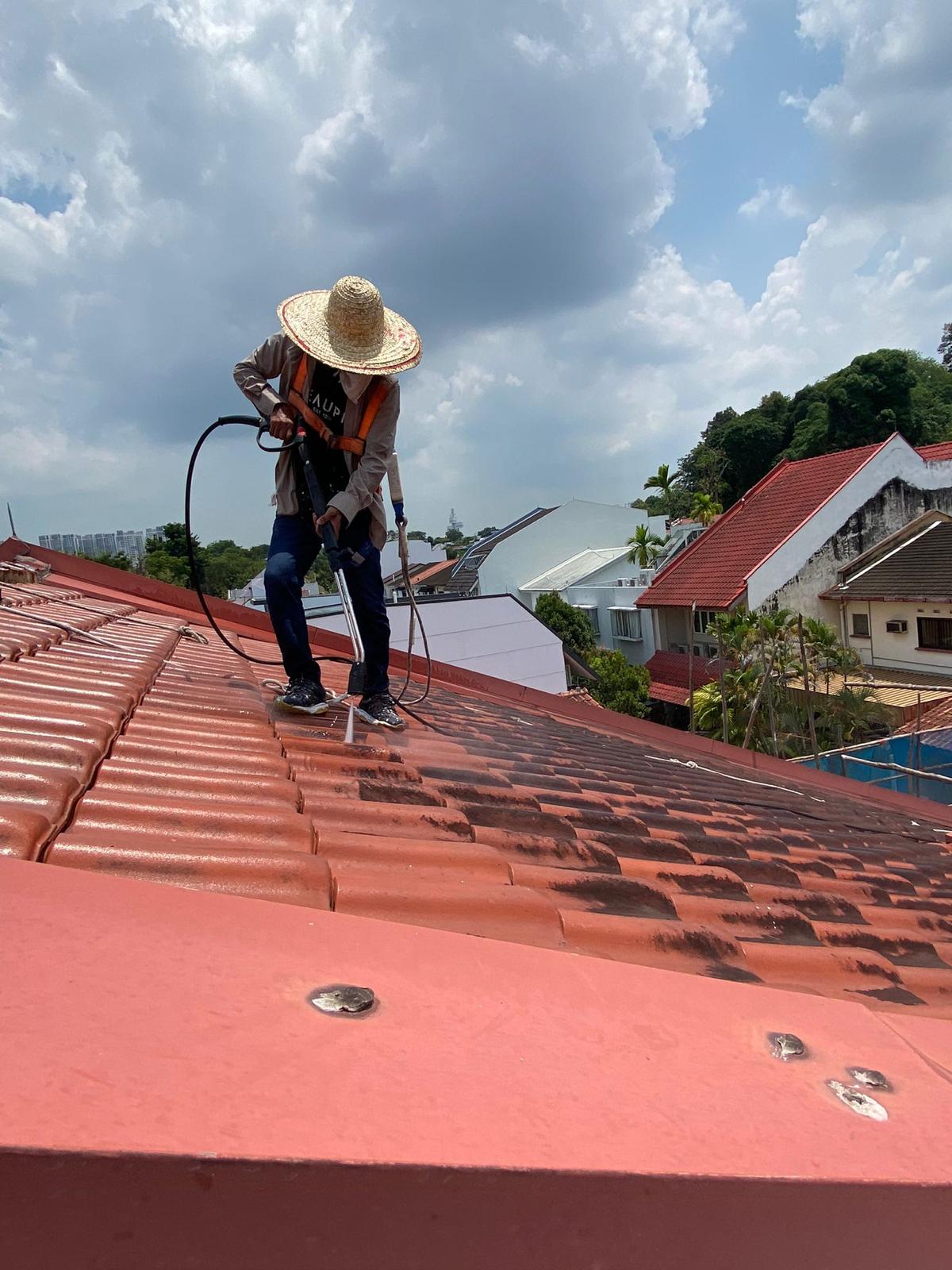Person wearing a straw hat power washing dirt off a red tiled roof under cloudy sky.