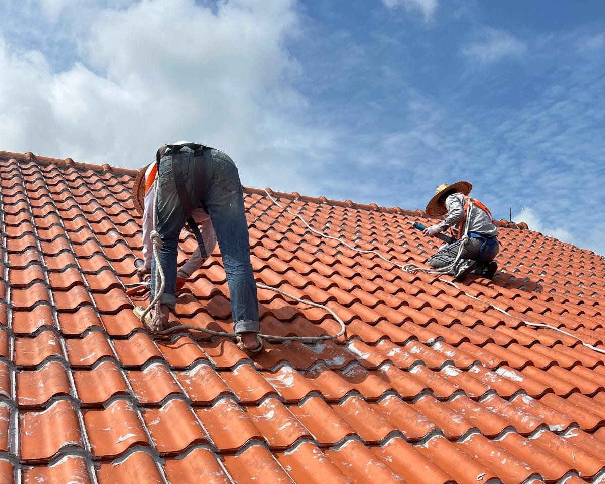 Two workers installing or repairing an orange clay tile roof under a partly cloudy blue sky.