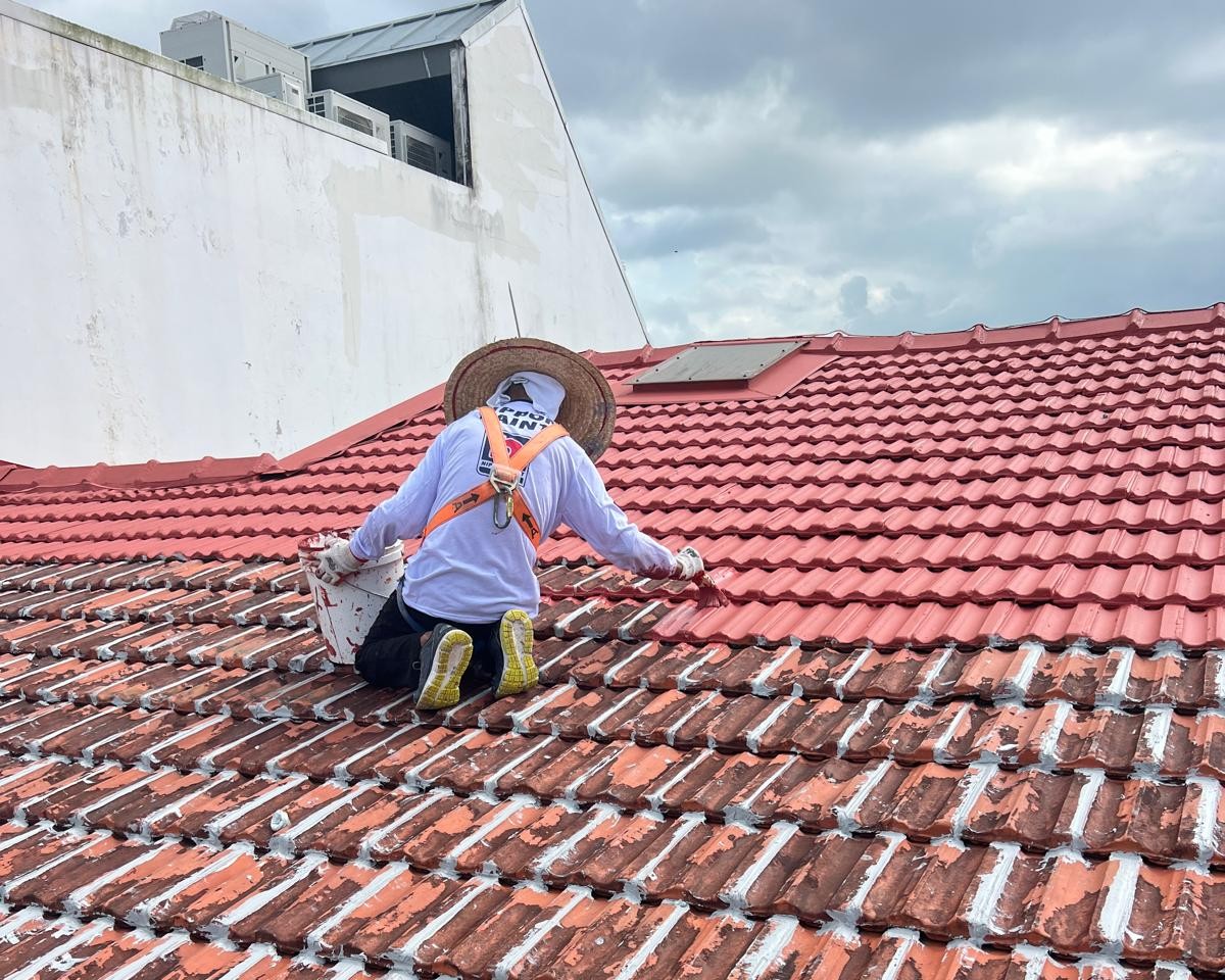 Person in safety harness painting clay roof tiles red on a cloudy day.