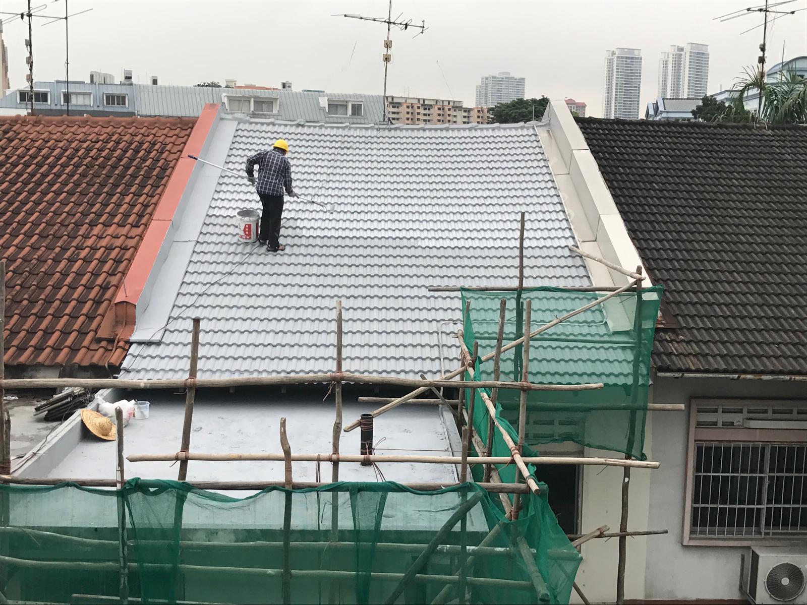 Worker in yellow helmet painting white tiles on a rooftop between brown and black tiled roofs.