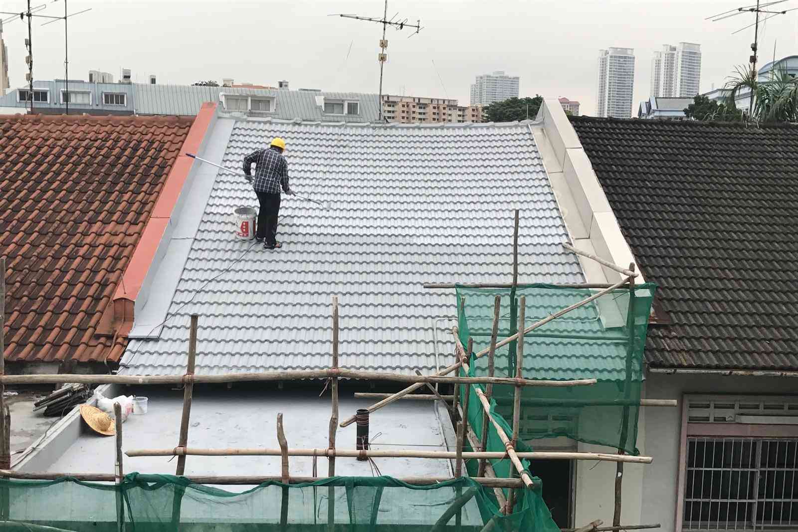 Worker in a yellow helmet painting a roof white while standing on a rooftop scaffold.
