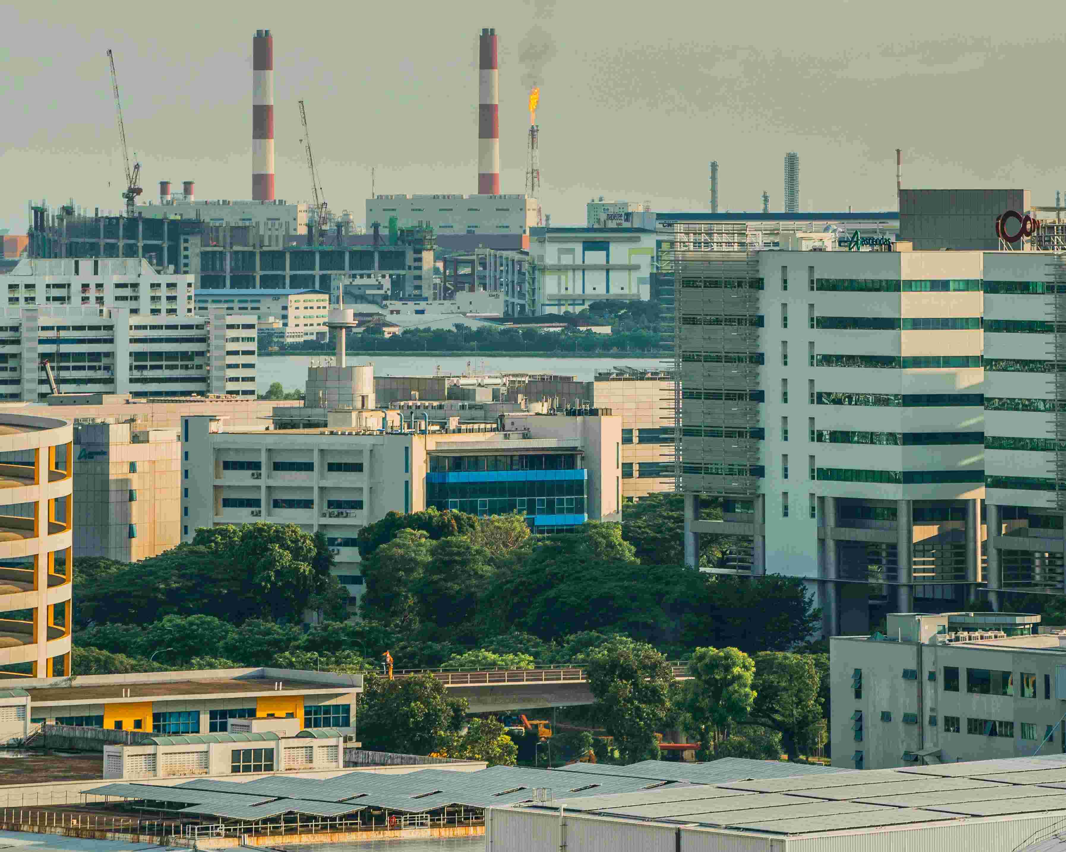 Urban industrial area with modern buildings, cranes, chimneys, and a gas flare against a hazy sky.