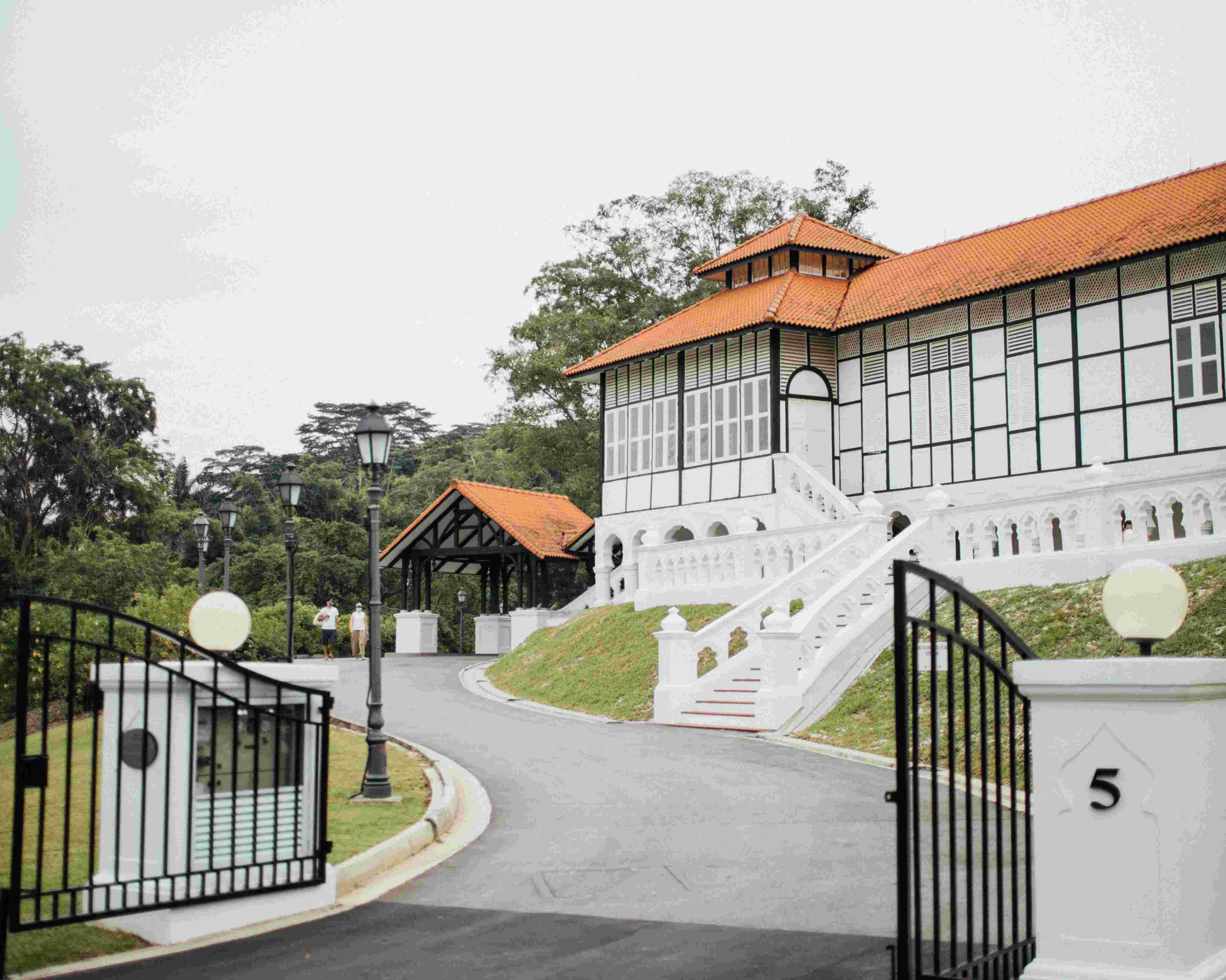 Large white bungalow with orange tiled roof and stairs, black iron gate, and two people walking on path.