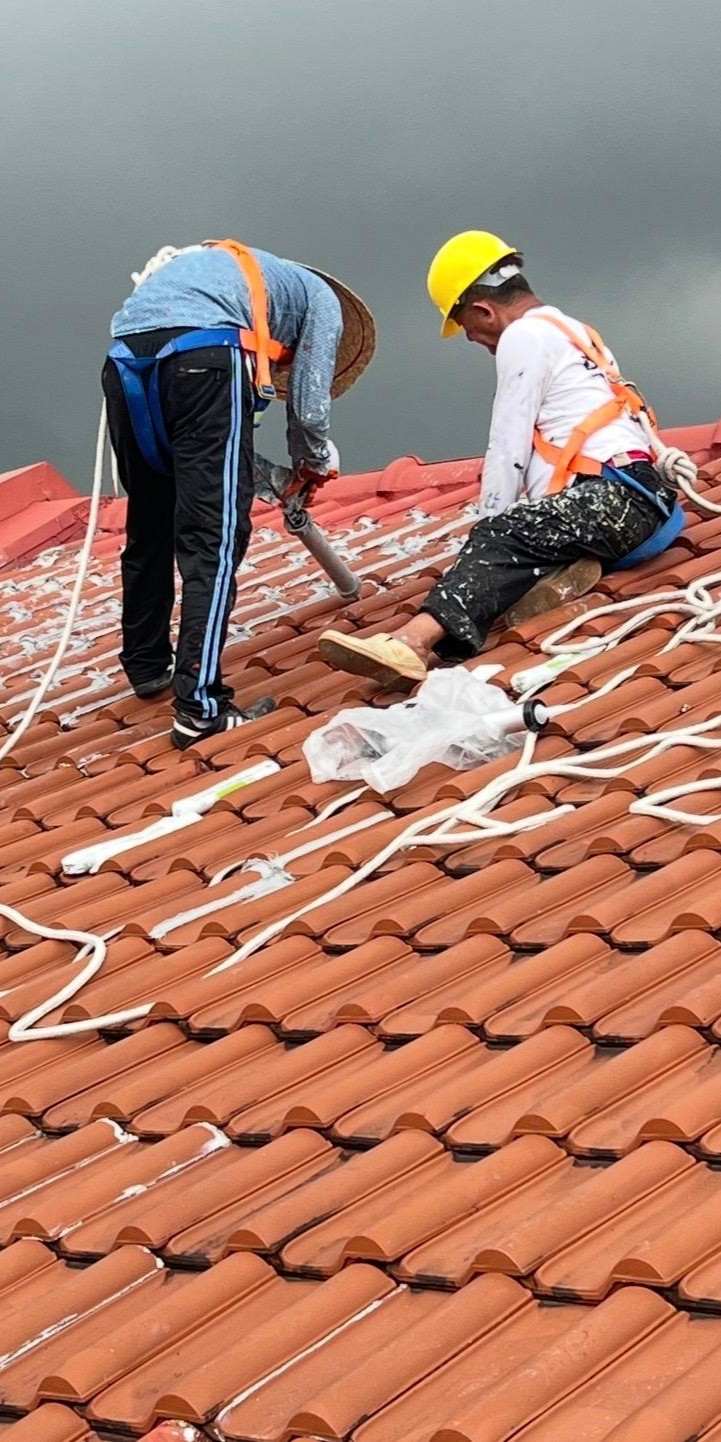 Two workers in safety harnesses applying sealant on a red tiled roof under a gray sky.