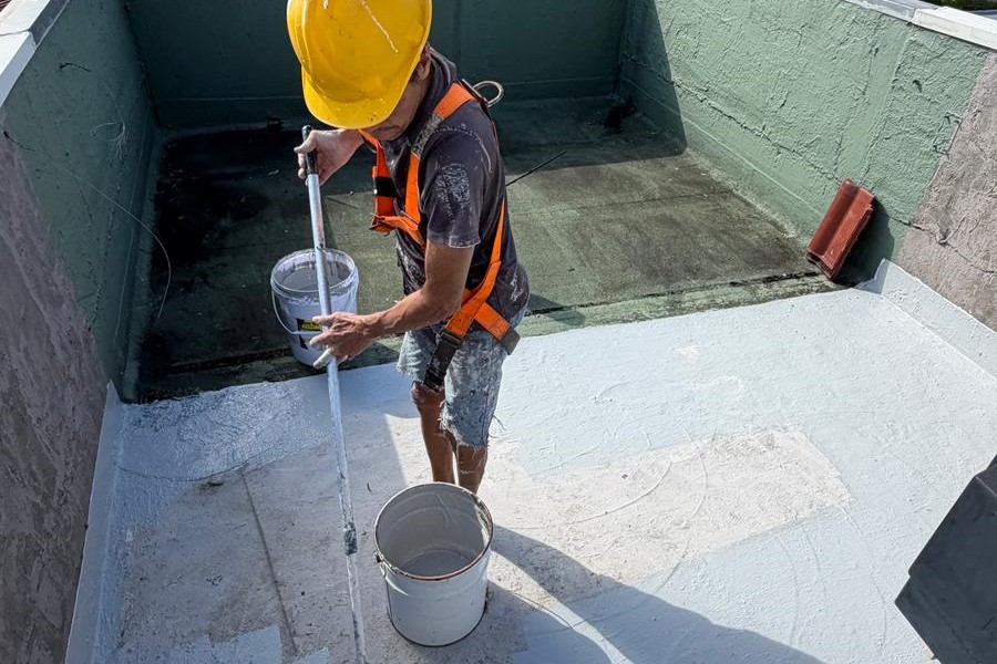 Worker wearing yellow helmet and safety harness painting a rooftop white with a roller.