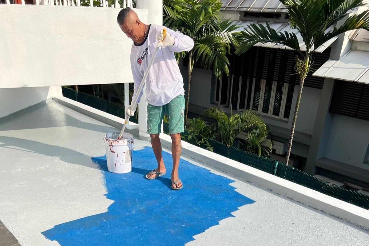 Man in white shirt painting a flat roof blue with a roller under sunlight.