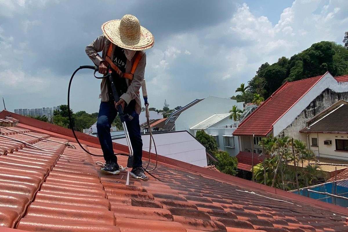 Person wearing a straw hat pressure washing red roof tiles on a sunny day with cloudy sky.